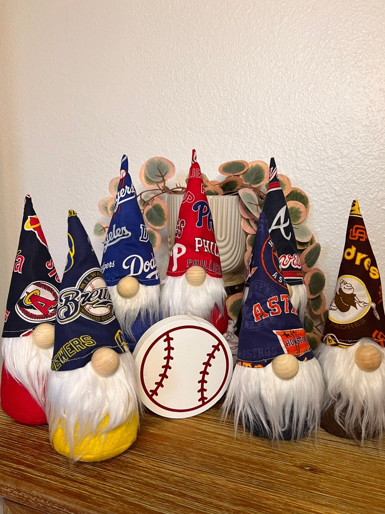 Puede incluir: Siete gnomos con logotipos de equipos de b&eacute;isbol en sus sombreros. Los gnomos visten ropa de diferentes colores y tienen barbas blancas. Una pelota de b&eacute;isbol blanca est&aacute; en el centro de la imagen.