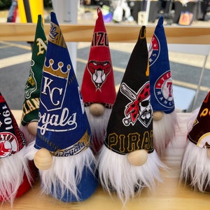 Puede incluir: Una colecci&oacute;n de siete adornos con forma de gnomo con diferentes logotipos de equipos de b&eacute;isbol. Los gnomos est&aacute;n hechos de tela y tienen barbas blancas. Los logotipos incluyen los Kansas City Royals, los Texas Rangers, los Pittsburgh Pirates, los Arizona Diamondbacks, los Boston Red Sox y dos m&aacute;s.