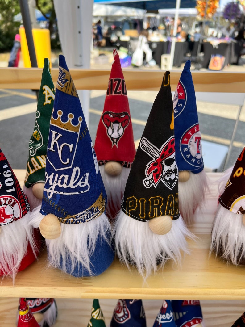 Puede incluir: Una colecci&oacute;n de siete adornos con forma de gnomo con diferentes logotipos de equipos de b&eacute;isbol. Los gnomos est&aacute;n hechos de tela y tienen barbas blancas. Los logotipos incluyen los Kansas City Royals, los Texas Rangers, los Pittsburgh Pirates, los Arizona Diamondbacks, los Boston Red Sox y dos m&aacute;s.