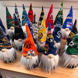 May include: A collection of 14 gnomes with different NHL team logos on their hats. The gnomes are arranged in a row on a wooden surface. The logos include the Vegas Golden Knights, Edmonton Oilers, Florida Panthers, New York Rangers, and more.