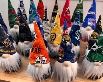 May include: A collection of 14 gnomes with different NHL team logos on their hats. The gnomes are arranged in a row on a wooden surface. The logos include the Vegas Golden Knights, Edmonton Oilers, Florida Panthers, New York Rangers, and more.