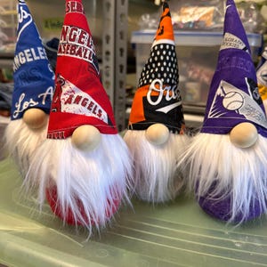 Puede incluir: Tres gnomos con barbas blancas y sombreros rojos, naranja y morados. Los sombreros est&aacute;n decorados con logotipos de equipos de b&eacute;isbol, incluidos los Los Angeles Angels, los Oakland Athletics y los Colorado Rockies.