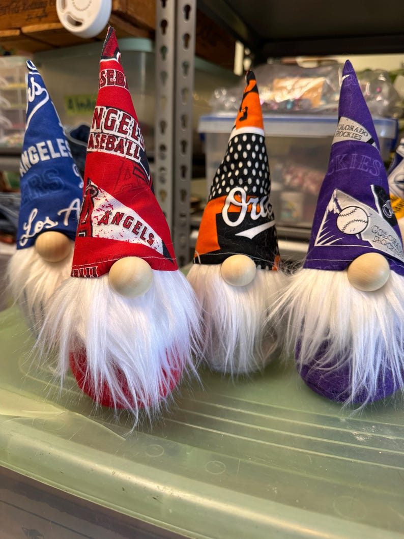 Puede incluir: Tres gnomos con barbas blancas y sombreros rojos, naranja y morados. Los sombreros est&aacute;n decorados con logotipos de equipos de b&eacute;isbol, incluidos los Los Angeles Angels, los Oakland Athletics y los Colorado Rockies.