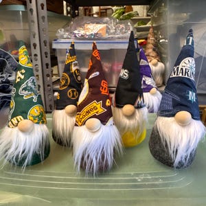 Puede incluir: Seis gnomos con barbas blancas y diferentes logotipos de equipos de b&eacute;isbol en sus sombreros. Los sombreros son verdes, negros, marrones, azules, morados y negros con letras blancas. Los gnomos est&aacute;n sentados sobre una superficie verde.
