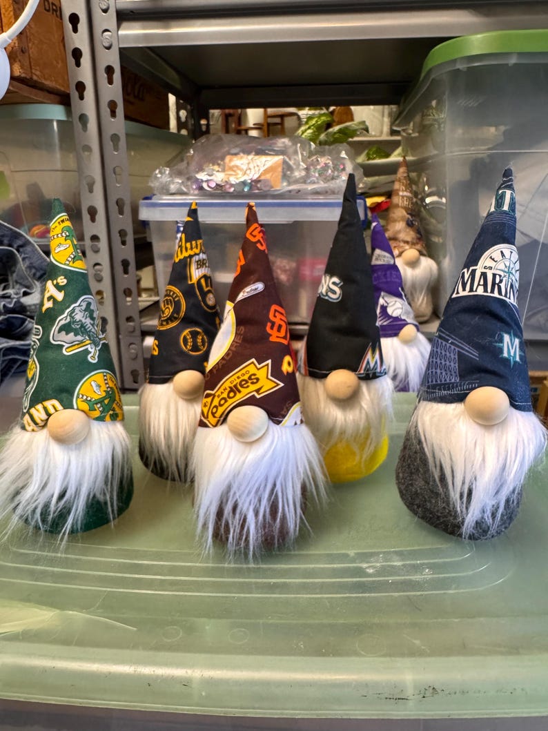 Puede incluir: Seis gnomos con barbas blancas y diferentes logotipos de equipos de b&eacute;isbol en sus sombreros. Los sombreros son verdes, negros, marrones, azules, morados y negros con letras blancas. Los gnomos est&aacute;n sentados sobre una superficie verde.