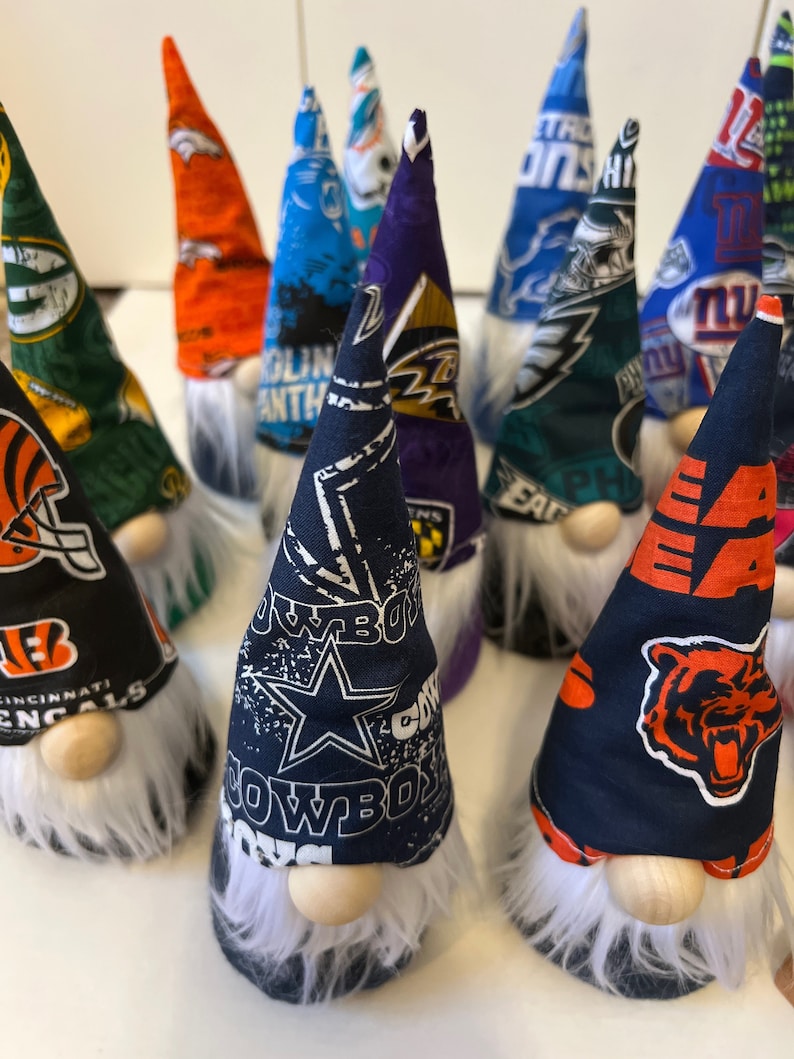 May include: A collection of handmade gnomes featuring various NFL team logos. The gnomes are crafted from fabric and have white beards. The logos include the Cincinnati Bengals, Dallas Cowboys, Philadelphia Eagles, Baltimore Ravens, Chicago Bears, New York Giants, and Denver Broncos.