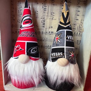 May include: Two fabric gnomes with tall pointed hats. The left gnome features red, black, and white Carolina Hurricanes team logos. The right gnome features black, grey, and gold Vegas Golden Knights team logos. Both have white faux fur beards.