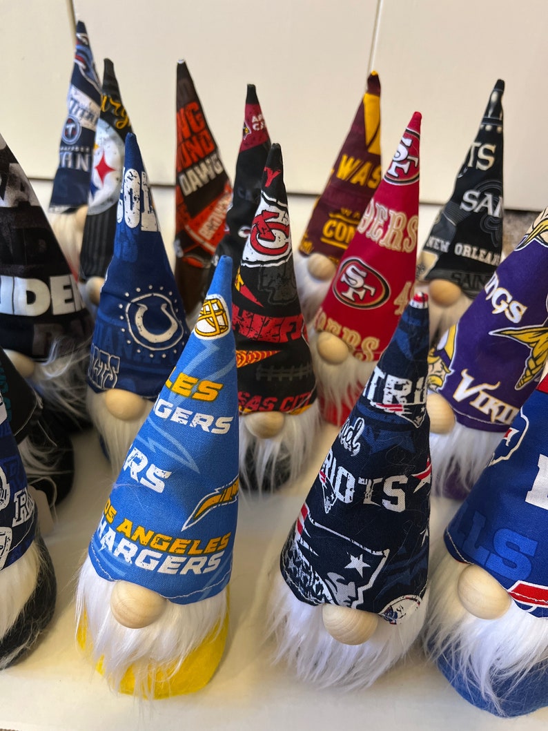 May include: A collection of handmade gnomes featuring various NFL team logos. The gnomes are crafted from fabric and have white fur beards. The logos include the Chargers, Patriots, Saints, Vikings, 49ers, and more.