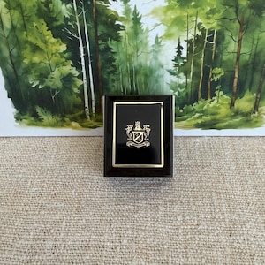 May include: A rectangular black jewelry box with a gold-toned border and a central gold crest emblem featuring a shield and decorative flourishes. The box has a polished finish and is positioned upright against a backdrop of a forest painting.
