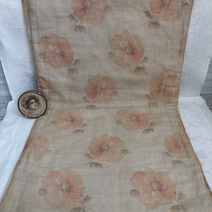 May include: A beige floral runner featuring a repeating pattern of large, peach-colored flowers with green leaves. The runner is displayed on a white surface, with a small framed portrait in the corner.