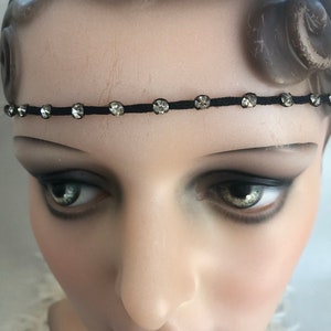 May include: A black headband with sparkling, clear rhinestones evenly spaced along its length. The headband is thin and delicate, designed to be worn across the forehead. The rhinestones catch the light, adding a touch of glamour.