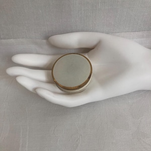 May include: A small, round, vintage trinket box with a pale green top and a gold-colored rim. The box has a white band around the middle. The box is held in a white hand.