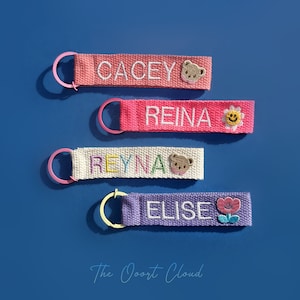 May include: Four personalized keychains with embroidered names and decorative elements. The keychains are in shades of pink, white, and purple, each with a different name: "Cacey", "Reina", "Reyna", and "Elise". The keychains have metal rings and are set against a blue background.