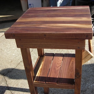 May include: A handcrafted wooden side table with a square top and a lower shelf. The table is made of dark brown wood with visible grain patterns. The table measures approximately 24 inches tall.