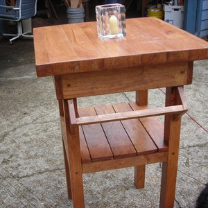 May include: A handcrafted wooden side table with a warm, reddish-brown finish. The table features a square top, a lower shelf, and sturdy legs. A clear glass candle holder with a lit candle sits on top. The table measures approximately 24 inches tall.