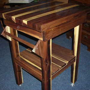 May include: A wooden kitchen cart with a butcher block top, a lower shelf, and a towel rack. The cart is made of dark wood with light wood accents and has wheels.