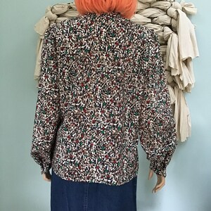 May include: A long-sleeved blouse featuring a dense, multi-colored geometric pattern of small triangles in shades of brown, white, black, and green. The blouse has a high neckline, full sleeves with buttoned cuffs, and is paired with a blue denim skirt.