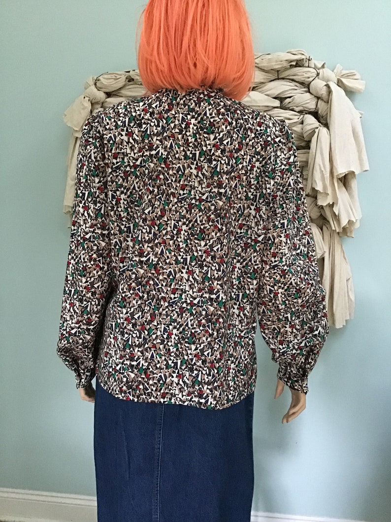 May include: A long-sleeved blouse featuring a dense, multi-colored geometric pattern of small triangles in shades of brown, white, black, and green. The blouse has a high neckline, full sleeves with buttoned cuffs, and is paired with a blue denim skirt.