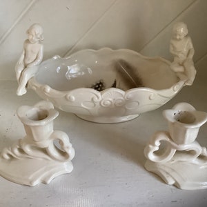 Può includere: Un centrotavola in ceramica color crema con due figure di cherubini e due candelabri abbinati. La ciotola ha un bordo smerlato e dettagli decorativi. Ci sono delle piume all'interno della ciotola.