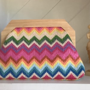 May include: A colorful, chevron-patterned clutch with a wooden frame. The bag features a vibrant mix of pink, orange, yellow, green, blue, and white. A small, cream-colored statue is to the right of the bag.
