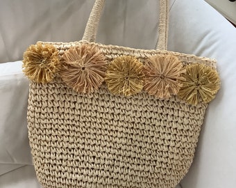 May include: Woven straw tote bag with a rounded shape and sturdy handles. The top edge is adorned with five decorative, flower-like accents in shades of tan and beige. The bag is designed for carrying items.