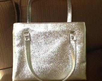 May include: Silver tote bag with a textured surface and two handles. The bag has a rectangular shape with a top handle and two smaller handles attached to the sides. The bag is made of a shiny, metallic material.
