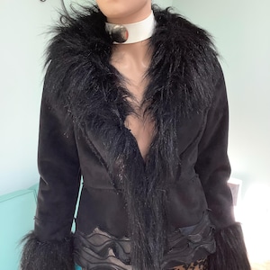 May include: A black jacket featuring a wide faux fur collar and matching faux fur cuffs on the long sleeves. The jacket has a smooth matte finish and a decorative wavy layered trim along the bottom hemline, worn over a leopard print skirt.