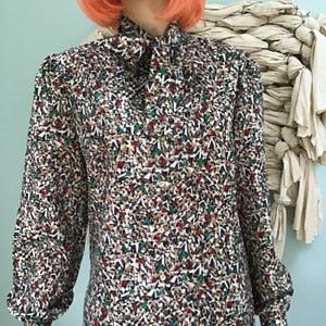 May include: A long-sleeved button-down blouse featuring a high neck with a tie detail. The fabric displays a dense, multi-colored geometric pattern consisting of small triangles in shades of brown, white, dark blue, green, and red throughout.