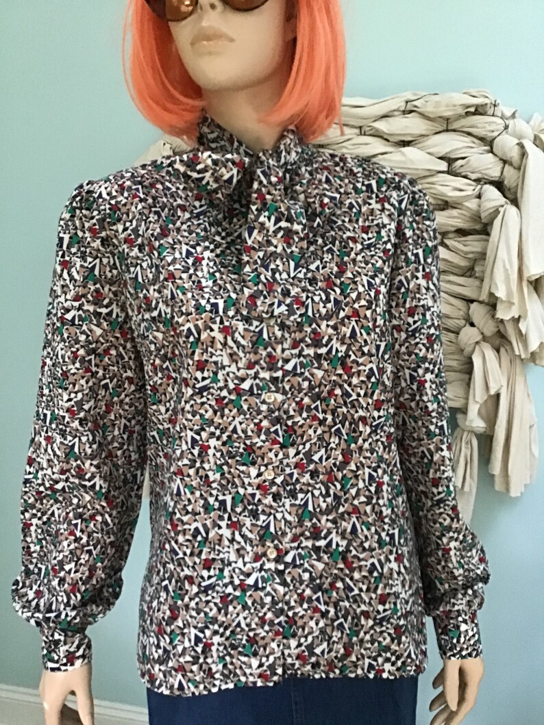 May include: A long-sleeved button-down blouse featuring a high neck with a tie detail. The fabric displays a dense, multi-colored geometric pattern consisting of small triangles in shades of brown, white, dark blue, green, and red throughout.