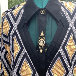 May include: A long-sleeved shirt featuring a black and gold animal print pattern with white geometric Greek key borders. The shirt has a solid black collar, a central black placket with a gold-toned button, and a decorative gold-toned brooch.
