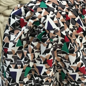 May include: A blouse featuring a gathered puff sleeve and a geometric pattern composed of white, black, tan, red, green, and dark blue triangular and abstract shapes. The fabric has a smooth texture and the sleeve is set into a structured shoulder.