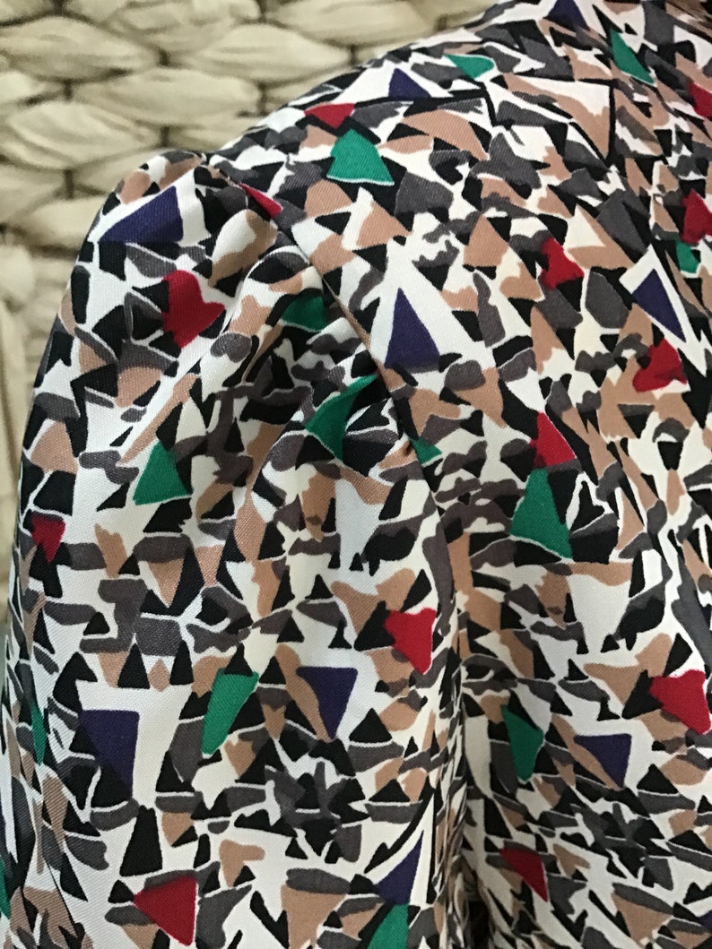 May include: A blouse featuring a gathered puff sleeve and a geometric pattern composed of white, black, tan, red, green, and dark blue triangular and abstract shapes. The fabric has a smooth texture and the sleeve is set into a structured shoulder.