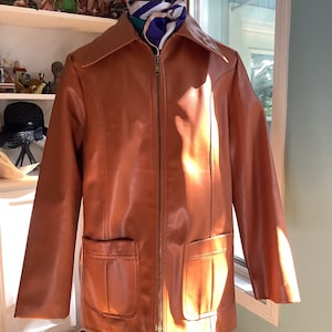 May include: A vintage, long-sleeved, brown leather jacket with a zipper closure and two front pockets. The jacket is displayed on a mannequin, with a scarf around the neck. The jacket has a wide collar.