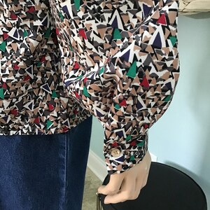 May include: A long-sleeved button-down shirt featuring a geometric pattern of triangles in shades of tan, black, white, red, green, and dark blue. The shirt has a structured cuff with a single light-colored button at the wrist and a collar.
