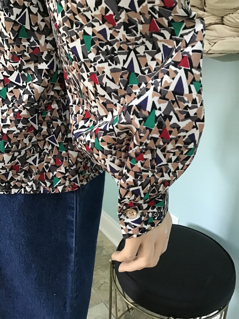 May include: A long-sleeved button-down shirt featuring a geometric pattern of triangles in shades of tan, black, white, red, green, and dark blue. The shirt has a structured cuff with a single light-colored button at the wrist and a collar.
