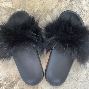 May include: A pair of black slide sandals with a fluffy, faux fur strap. The sandals have a solid black sole and a soft, textured fur band across the top. The image shows the sandals from a top-down perspective.