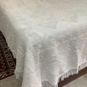 May include: A white bedspread featuring a textured, raised geometric pattern across the surface. The fabric has a distinct woven design with a decorative border and a fringe trim along the bottom edge, creating a tactile, monochromatic appearance.