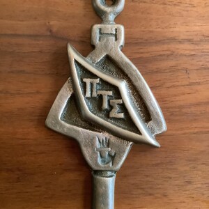 May include: A vintage metal key with a decorative design. The key features a loop at the top, a stylized shield with Greek letters, and a decorative element at the bottom. The key is made of a silver-colored metal and has a worn appearance.