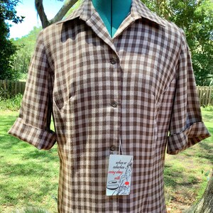 May include: A brown and white gingham button-down shirt with short sleeves and folded cuffs. The shirt features a pointed collar and a vertical row of buttons down the center. A white rectangular tag is attached, reading urban or suburban sewing along with Serben.