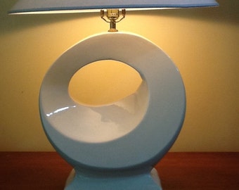 May include: A white ceramic table lamp with a circular design and a light blue lampshade. The lamp has a brass-colored base and a square base. The lampshade is triangular. The lamp is on a dark brown wooden surface.