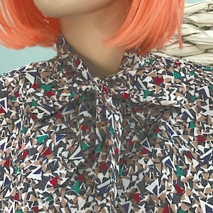 May include: A long-sleeved blouse featuring a high neck with a tied bow detail. The fabric displays a dense, geometric pattern composed of small, irregular triangular shapes in shades of dark gray, tan, white, deep red, dark blue, and bright green.