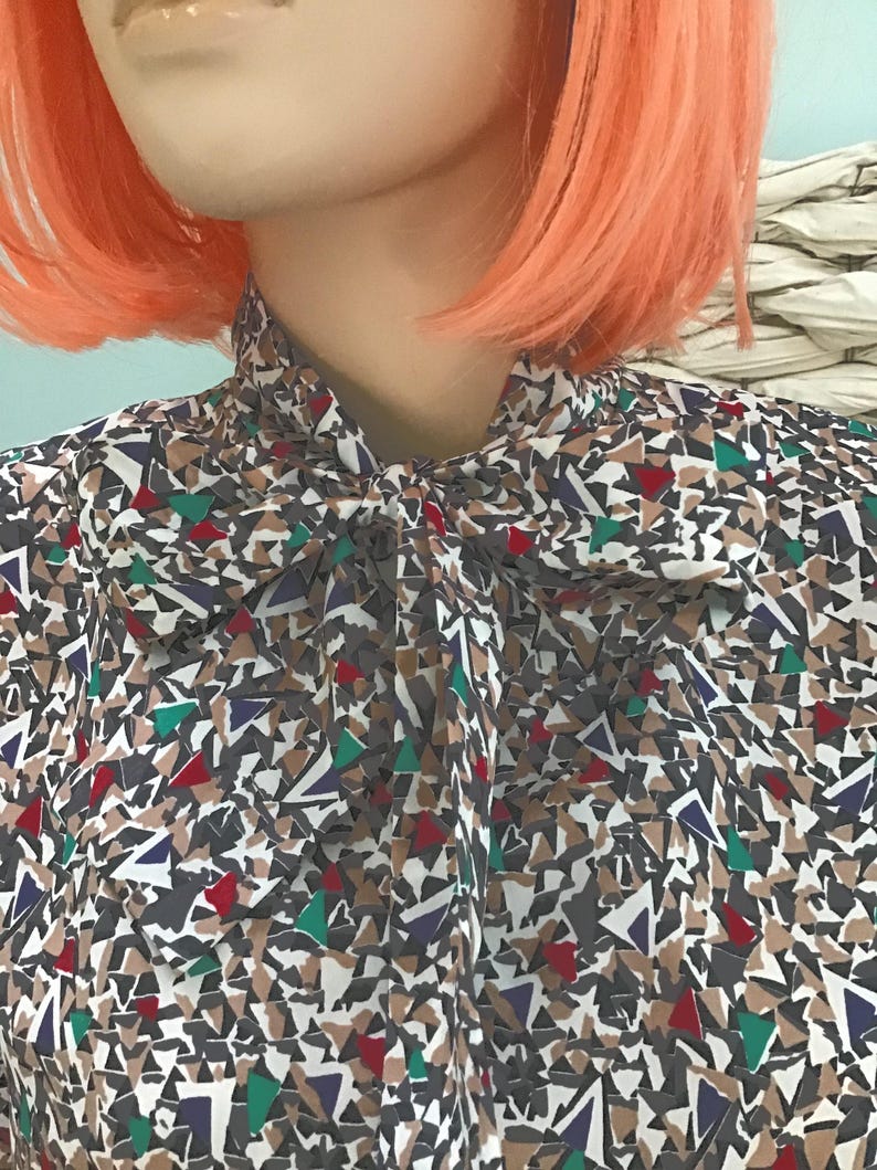 May include: A long-sleeved blouse featuring a high neck with a tied bow detail. The fabric displays a dense, geometric pattern composed of small, irregular triangular shapes in shades of dark gray, tan, white, deep red, dark blue, and bright green.
