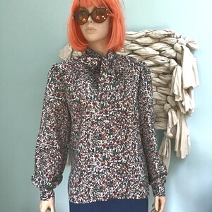 May include: A long-sleeved blouse featuring a high neck with a tie detail and a button-down front. The fabric displays a dense, multi-colored geometric pattern in shades of red, green, white, and brown. The sleeves are full with buttoned cuffs.