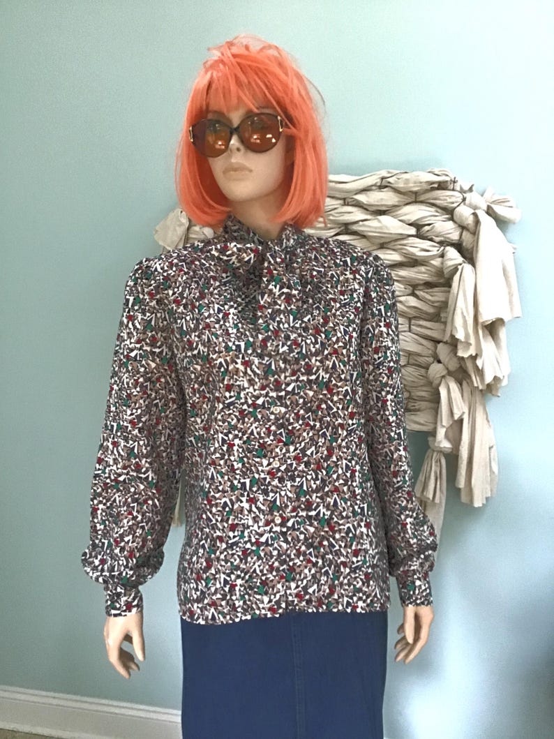 May include: A long-sleeved blouse featuring a high neck with a tie detail and a button-down front. The fabric displays a dense, multi-colored geometric pattern in shades of red, green, white, and brown. The sleeves are full with buttoned cuffs.