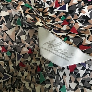 May include: A fabric garment featuring a geometric pattern of tan, gray, black, red, green, and purple triangular shapes. A white satin clothing label is sewn onto the fabric, displaying the brand name Alicia in a cursive script font.