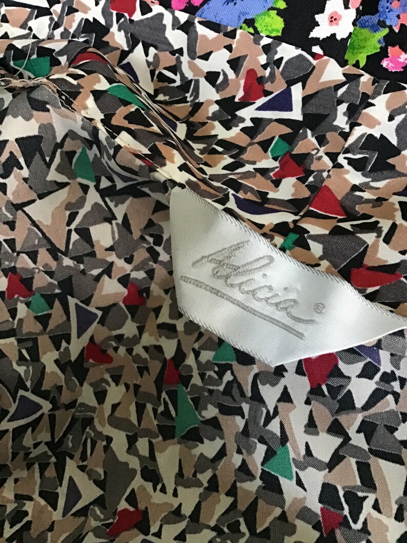 May include: A fabric garment featuring a geometric pattern of tan, gray, black, red, green, and purple triangular shapes. A white satin clothing label is sewn onto the fabric, displaying the brand name Alicia in a cursive script font.