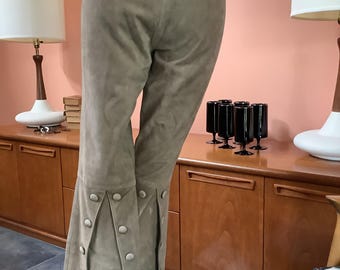 May include: Taupe suede pants with a high waist and flared leg. The pants feature decorative button detailing along the flared hem. The neutral color and classic design make these pants a versatile addition to any wardrobe.