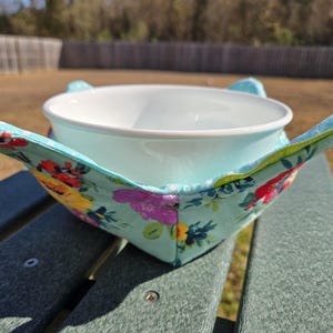 Puede incluir: Un cuenco blanco descansa dentro de un protector de cuenco con estampado floral. El protector es de color azul claro con un colorido estampado floral de flores rojas, amarillas y moradas. El protector tiene dos pequeñas asas en forma de oreja.