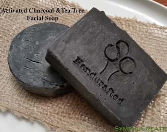 Puede incluir: Dos barras de jabón negras, una redonda y otra rectangular, con las palabras "Activated Charcoal & Tea Tree Facial Soap" y "Handcrafted" impresas en la barra rectangular.