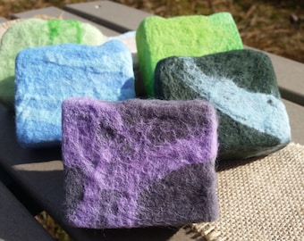 May include: Five handmade soap bars in various colours, including green, blue, purple, and grey. The soap bars are arranged in a row on a wooden surface.